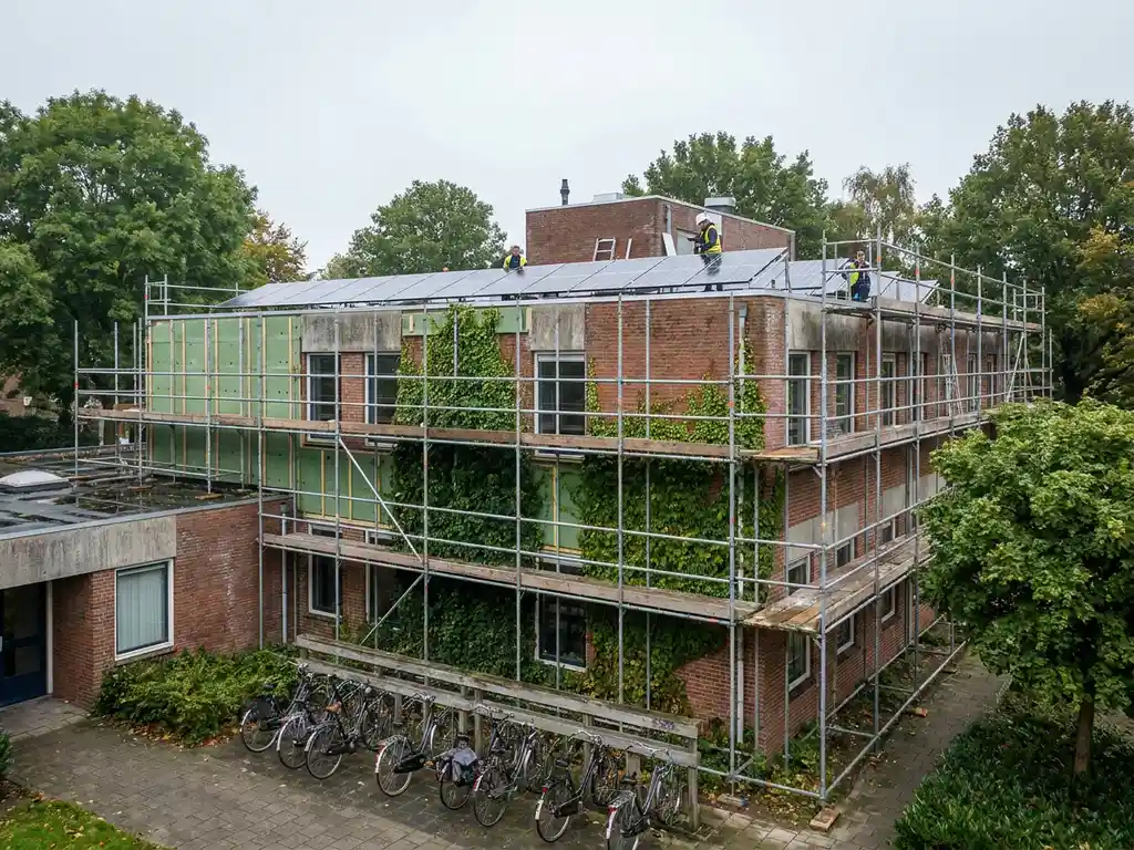 Kantoorgebouw in Nederland tijdens renovatie met zonnepanelen op dak, groene gevelisolatie en klimop op bakstenen gevel, omgeven door bomen en fietsenrek.