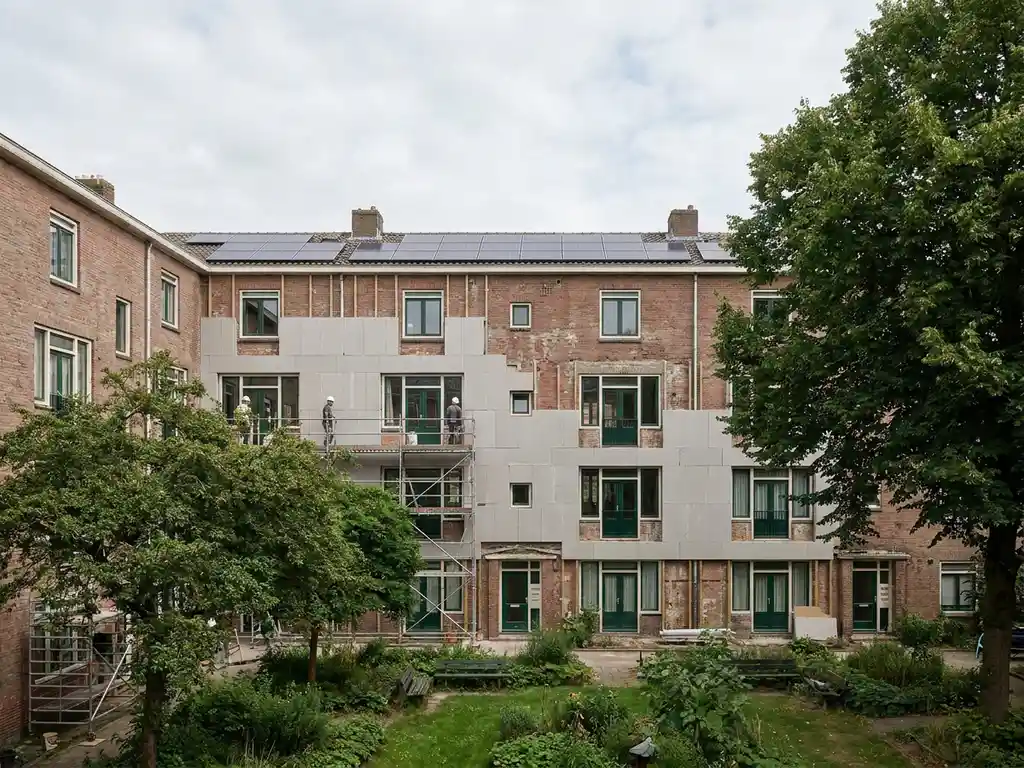 Sociale huurflat in renovatie met isolatiepanelen op bakstenen gevel en zonnepanelen op het dak, omgeven door groen.