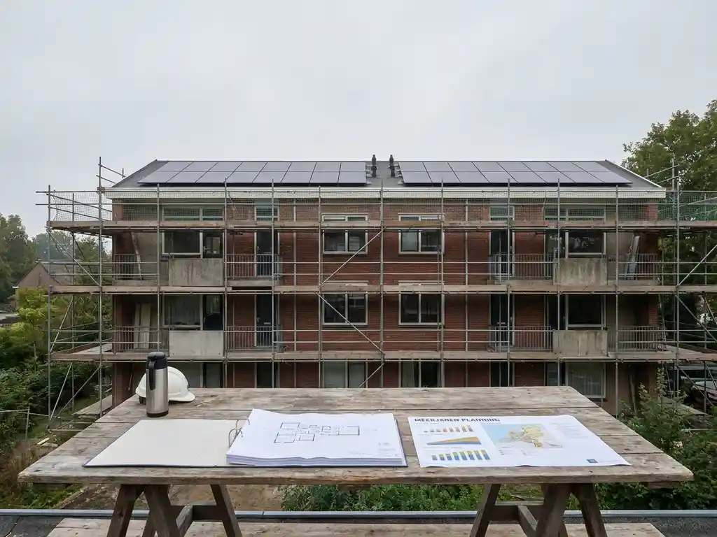 Sociale huurwoningen in renovatie met zonnepanelen op plat dak, steigers langs bakstenen gevel en opengeslagen planningsdocument op bouwplaatstafel.