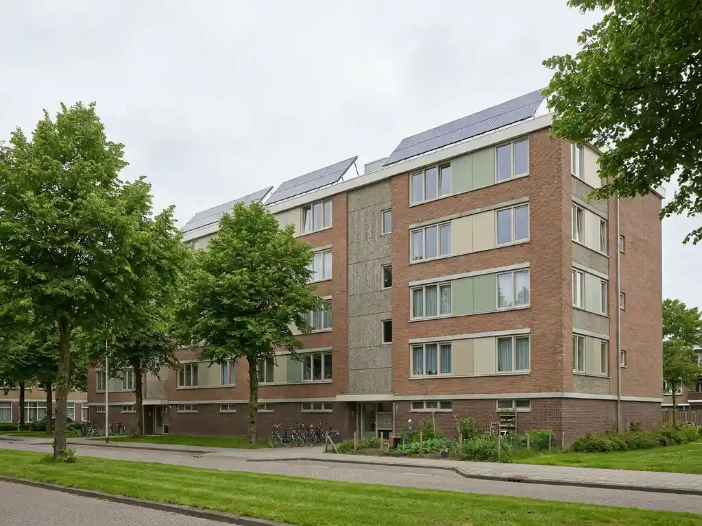 Gerenoveerd Nederlands sociale huurwoningblok met zonnepanelen en isolatiegevel, omgeven door volwassen bomen in een groene woonwijk.