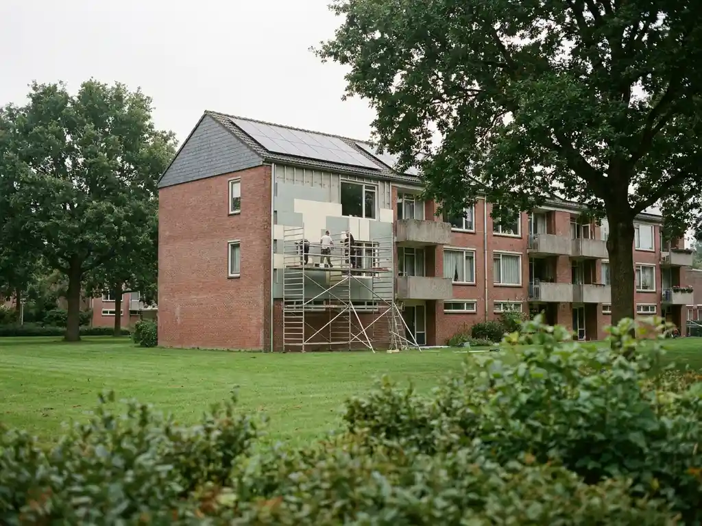 Sociale huurflat in renovatie met isolatiepanelen op bakstenen gevel, zonnepanelen op dak en steigers, omgeven door bomen.