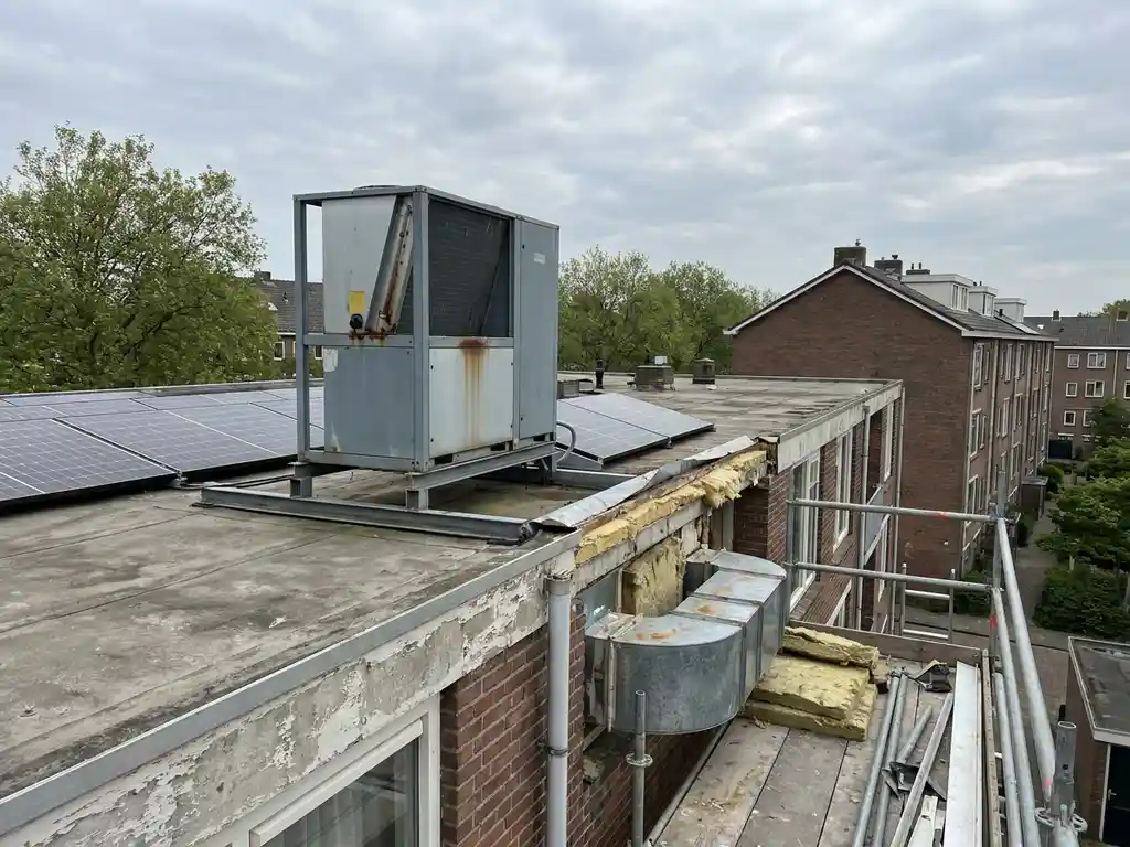 Warmtepomp op dak van Nederlandse sociale huurflat tijdens renovatie, met zonnepanelen, ventilatiekanalen en isolatielagen zichtbaar.