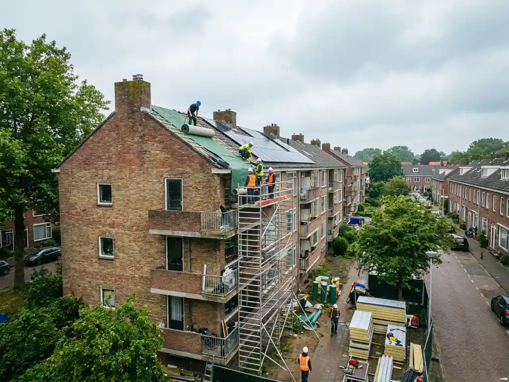 Arbeiders installeren zonnepanelen en groendakisolatie op een bakstenen appartementencomplex in een Nederlandse woonwijk.
