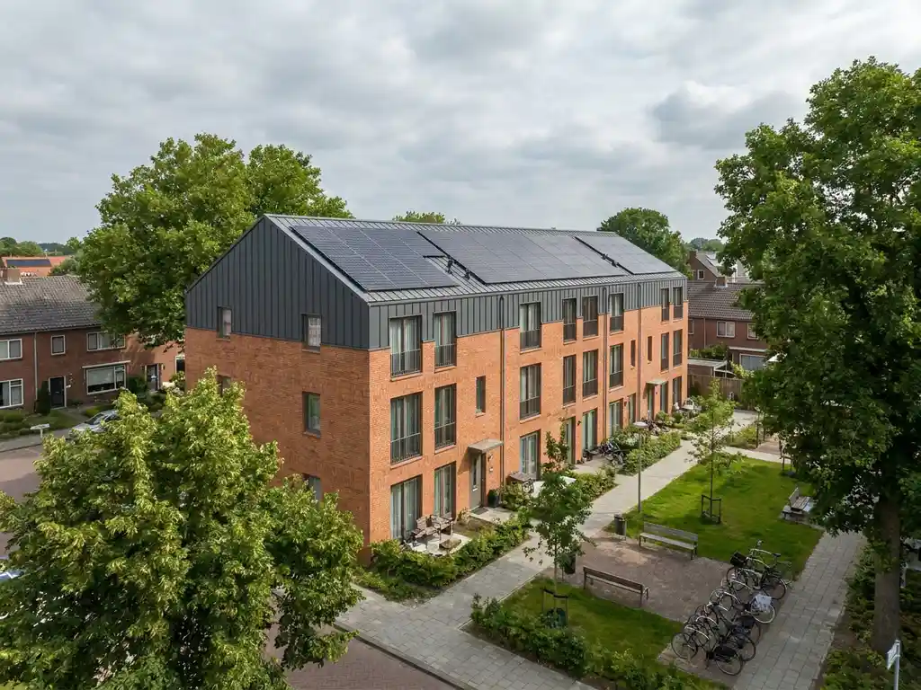 Nederlandstalige sociale huurflat met zonnepanelen op het volledige dak, omgeven door volwassen bomen en een verzorgde groene binnenplaats.