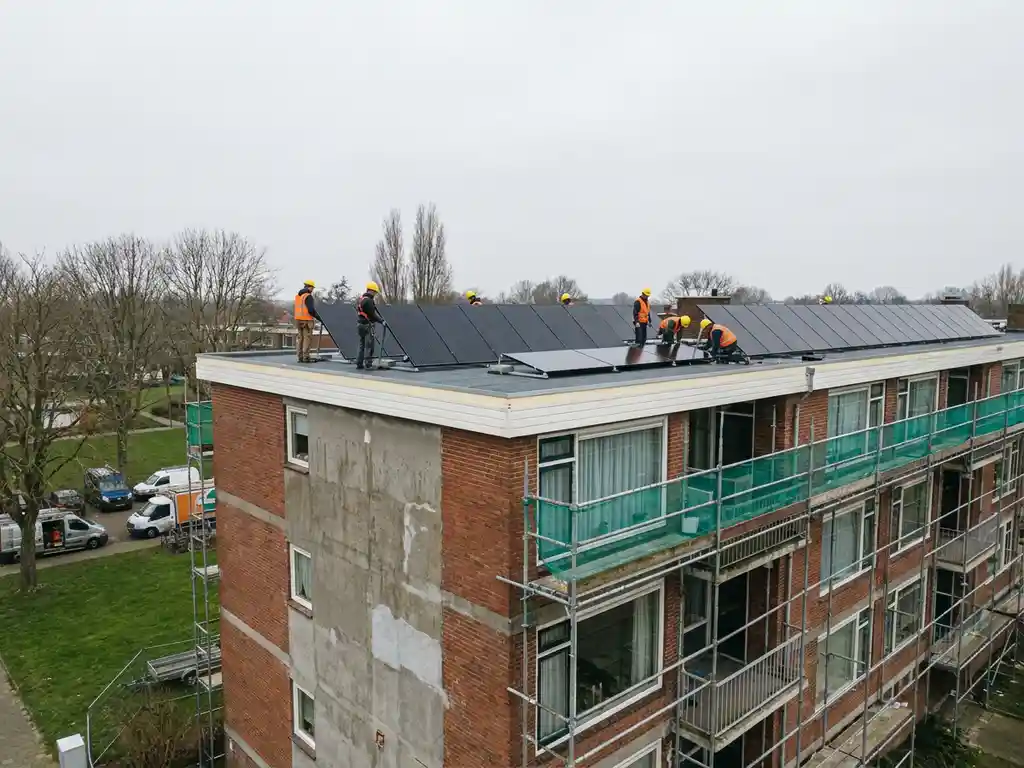 Arbeiders in veiligheidsuitrusting installeren zonnepanelen op geïsoleerd plat dak van Nederlandse sociale huurflat in renovatie.